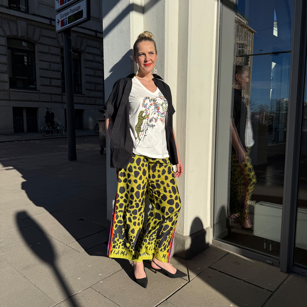 Frau steht in der Sonne vor einem Schaufenster, moderne Damenmode für große Größen in Münster mit auffälligen Farben und lockerem Stil.