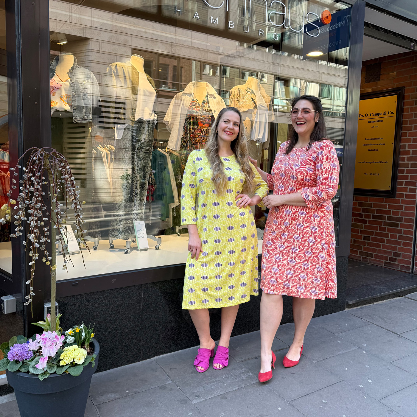 Zwei Frauen in farbigen Kleidern stehen vor einem Modegeschäft, elegante Karlsruhe Abendkleider große Größen mit femininen Schnitten und lebendigem Stil.