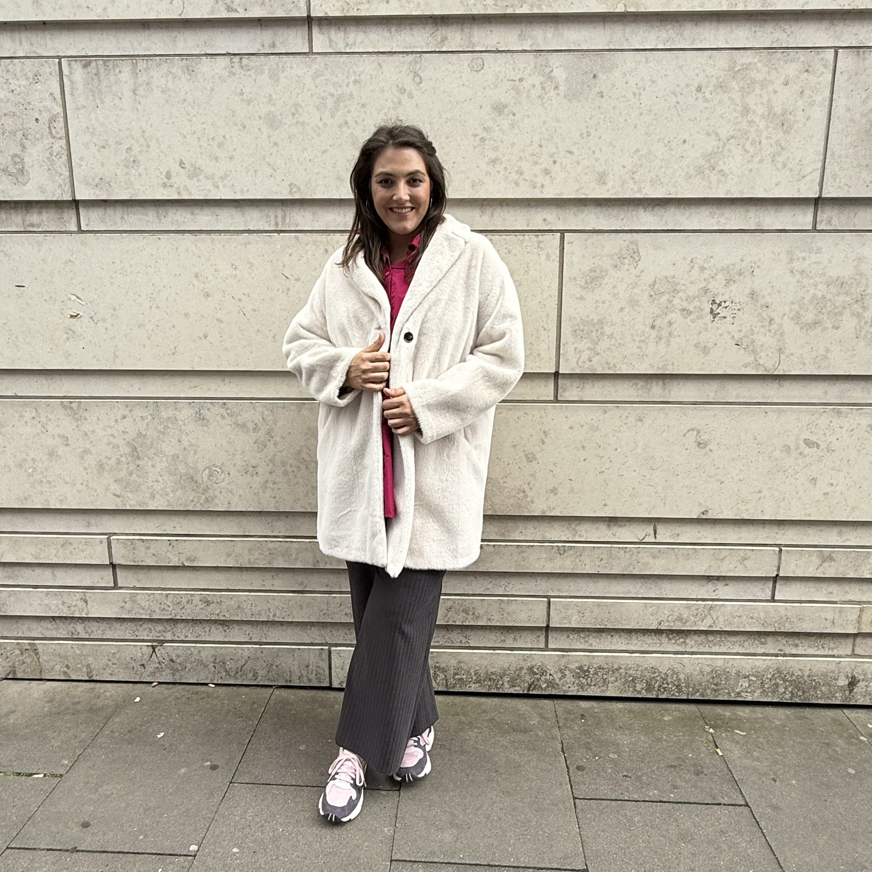 Frau trägt eleganten Mantel in Creme und weite Stoffhose vor einer Steinwand in Hannover, dezente Damenmode in großen Größen mit Stil.