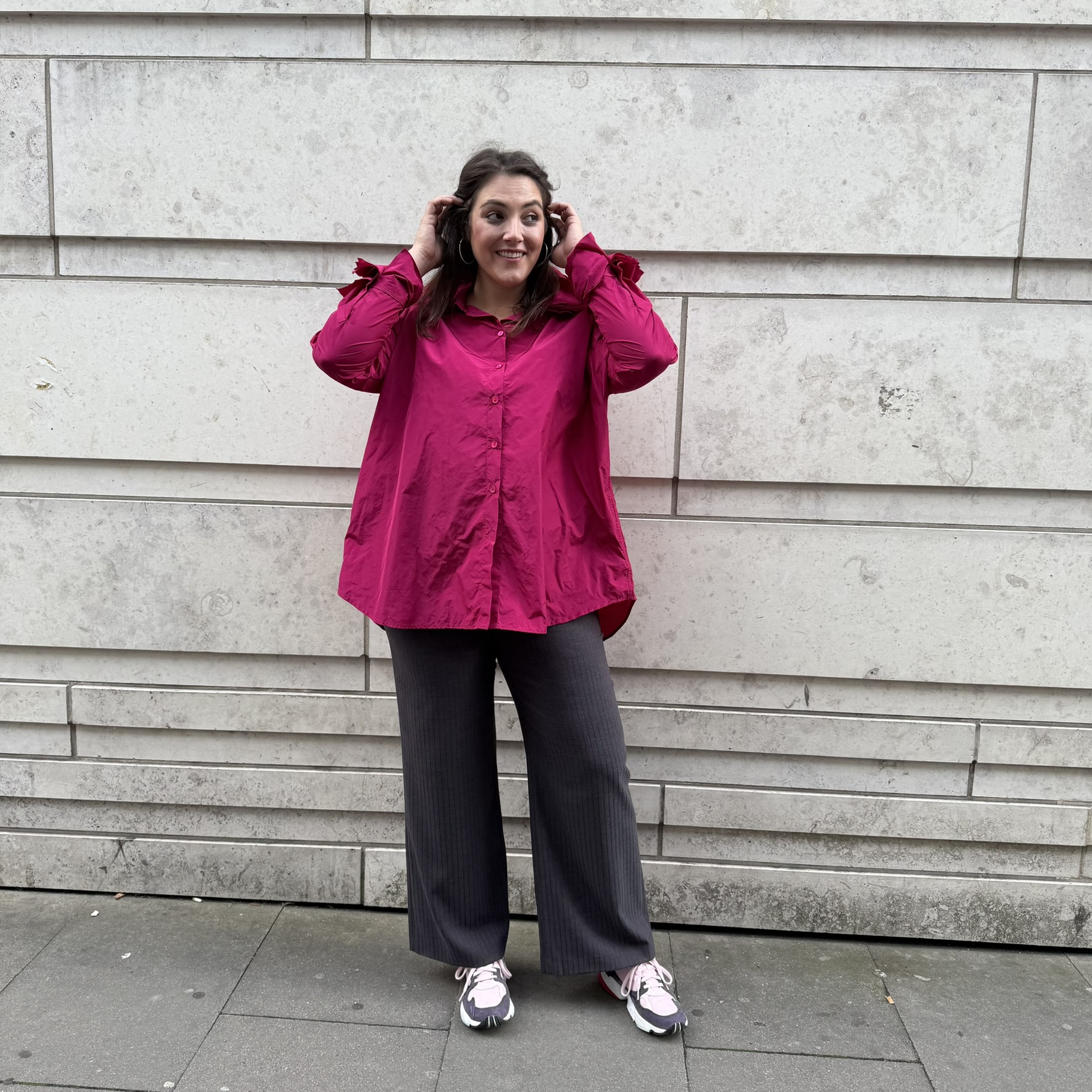 Frau trägt pinke Bluse und weite Stoffhose vor heller Steinwand, Mode für Mollige Damen München mit klarer Linie und bequemem Sitz.