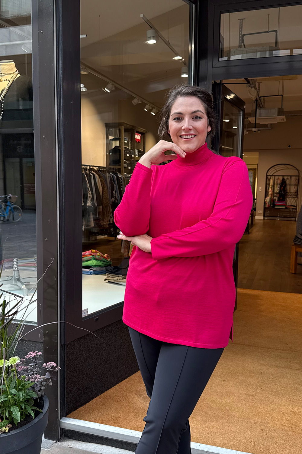Frau trägt pinken Pullover und dunkle Hose vor einer Boutique in Karlsruhe, moderne Plus Size Karlsruhe mit lässigem, stilvollem Auftritt.