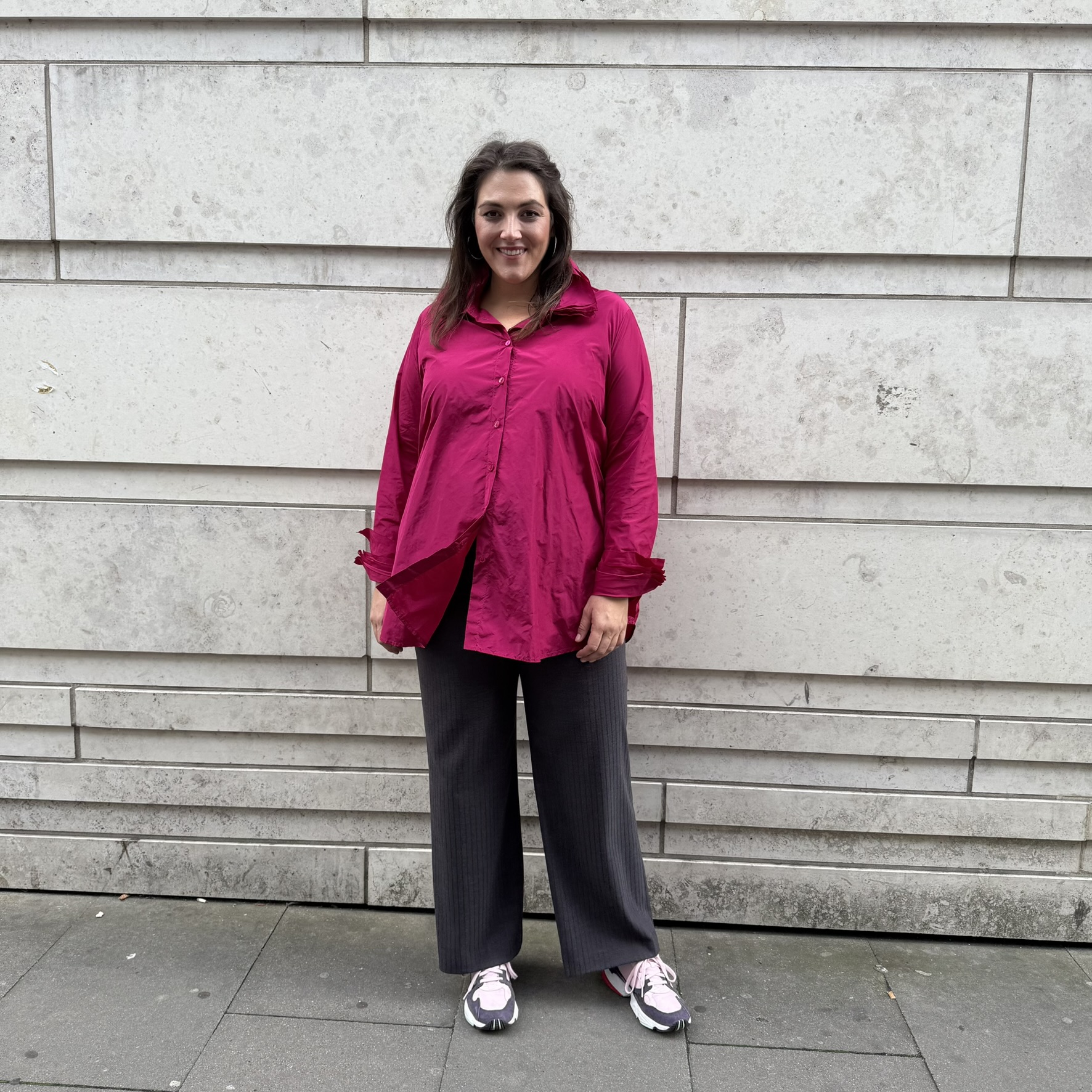 Frau trägt pinke Bluse und weite Stoffhose vor Steinwand in Berlin, moderne Plus Size Mode Berlin mit Stil und Selbstbewusstsein.