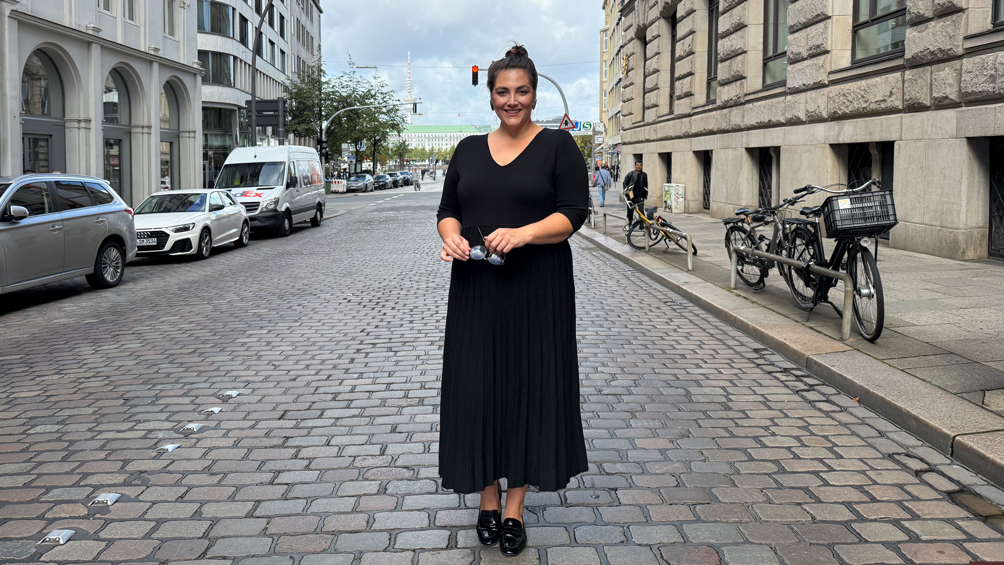 Frau in schwarzem Kleid steht auf einem gepflasterten Straßenabschnitt, elegante Übergrößen Münster Mode in klassischem, modernem Look.