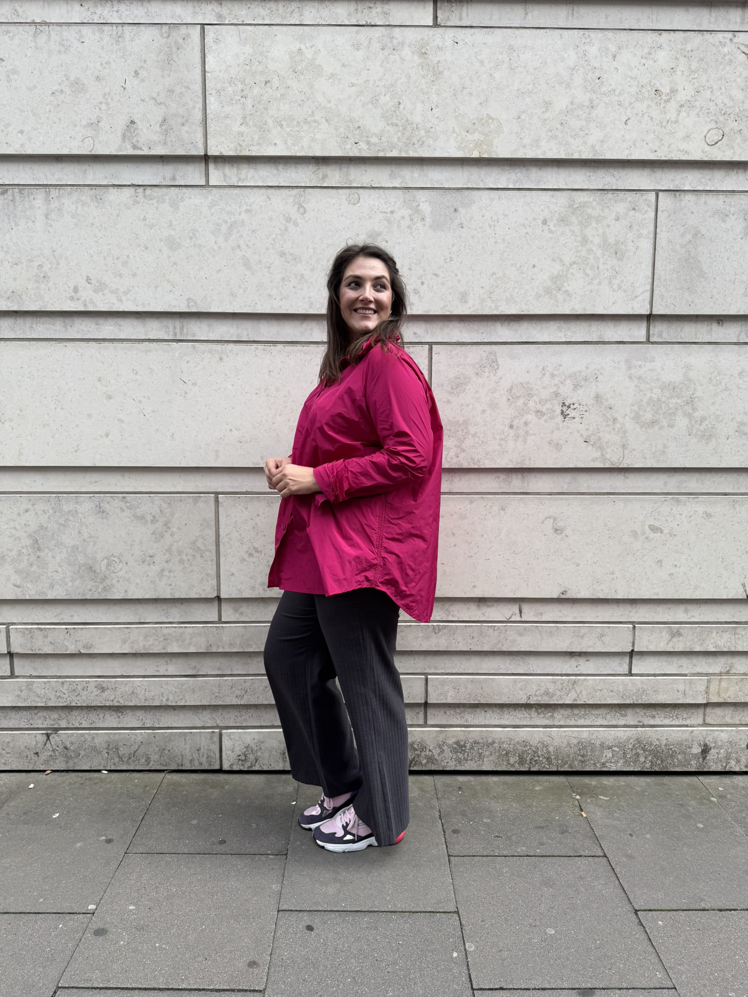Frau in pinker Bluse und dunkler Hose zeigt einen modernen Look für übergrößen leipzig vor einer hellen Steinwand.