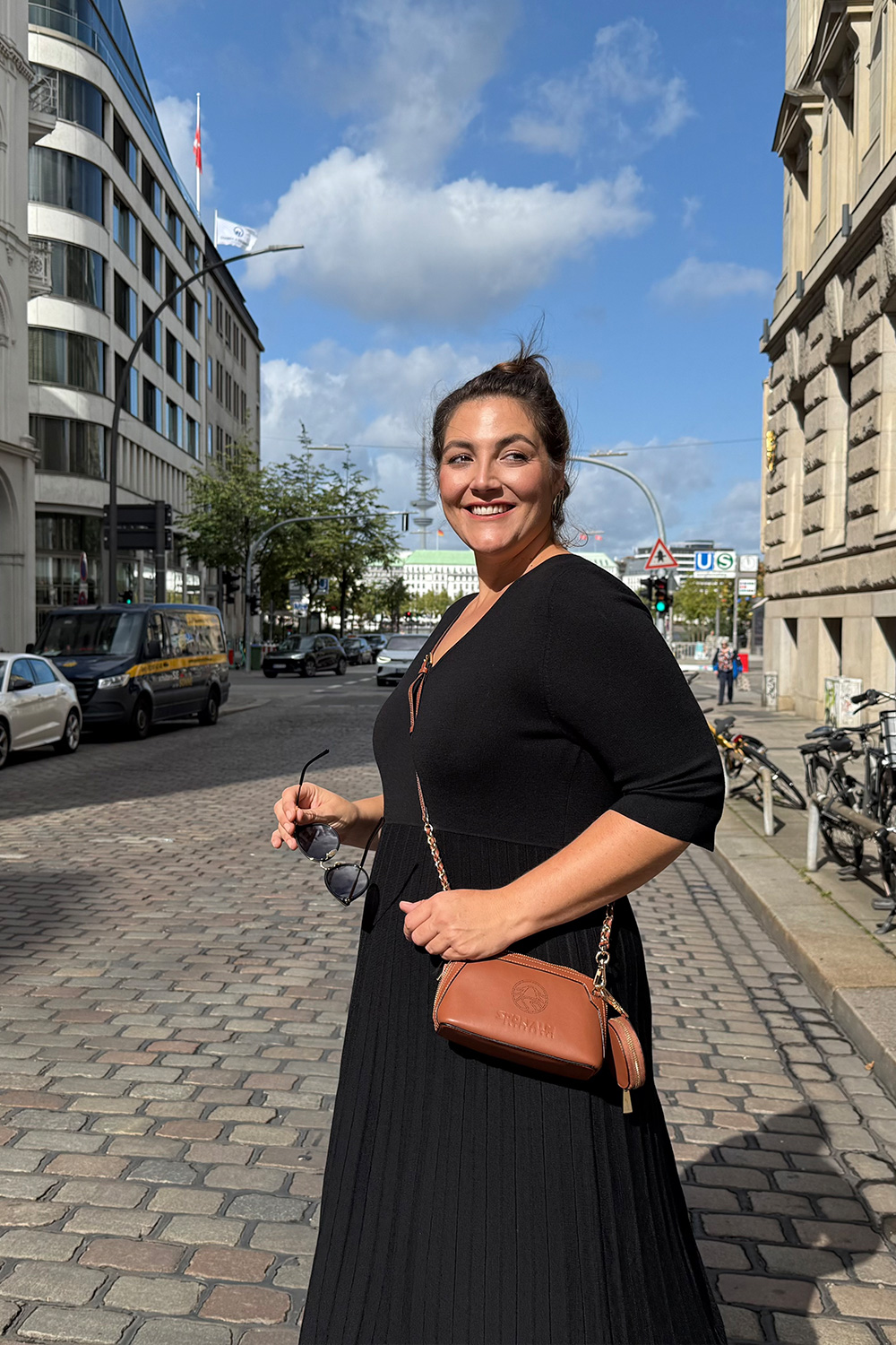 Abendkleider große Größen Braunschweig, Kundin in schwarzem Plisseekleid mit V Ausschnitt und cognacfarbener Umhängetasche.