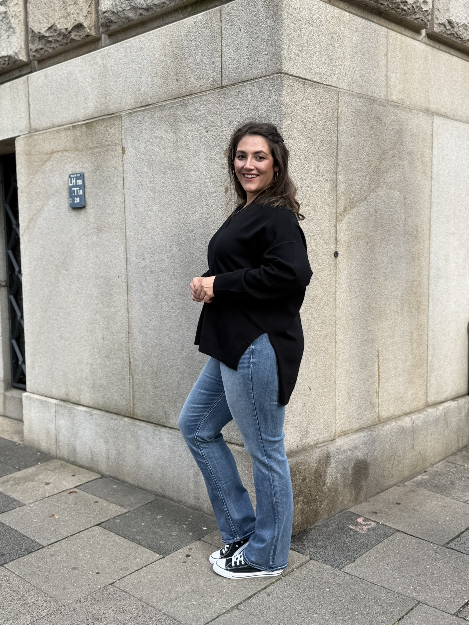 Übergrößen Mode Essen, Kundin in schwarzem Longpullover mit Seitenschlitzen, heller Bootcut Jeans und schwarzen Sneakern.