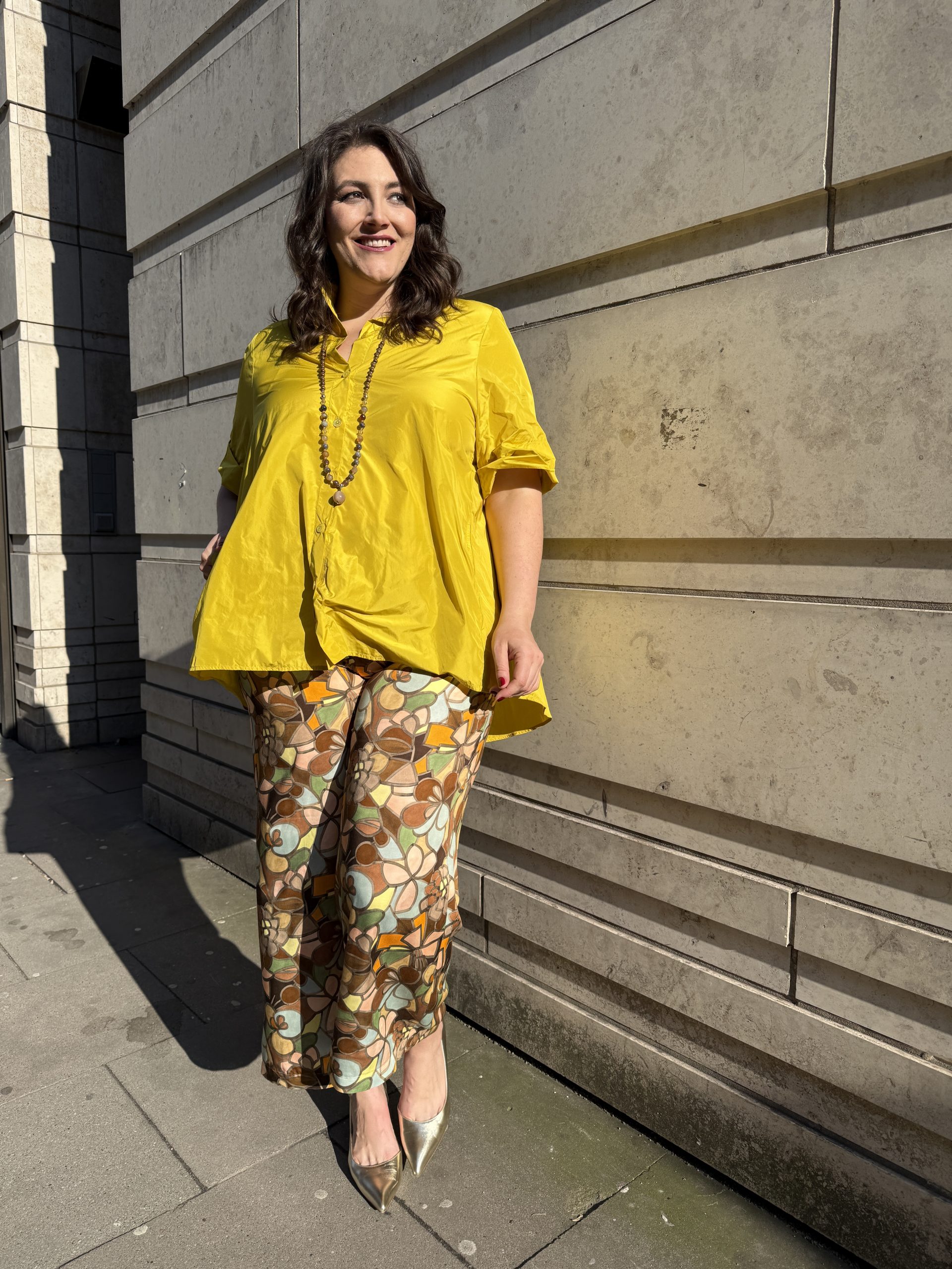 Lächelnde Kundin in gelber Seidenbluse und floral gemusterter Hose aus dem Kurvenhaus Hamburg, große Größen Augsburg in sommerlichem Streetstyle.