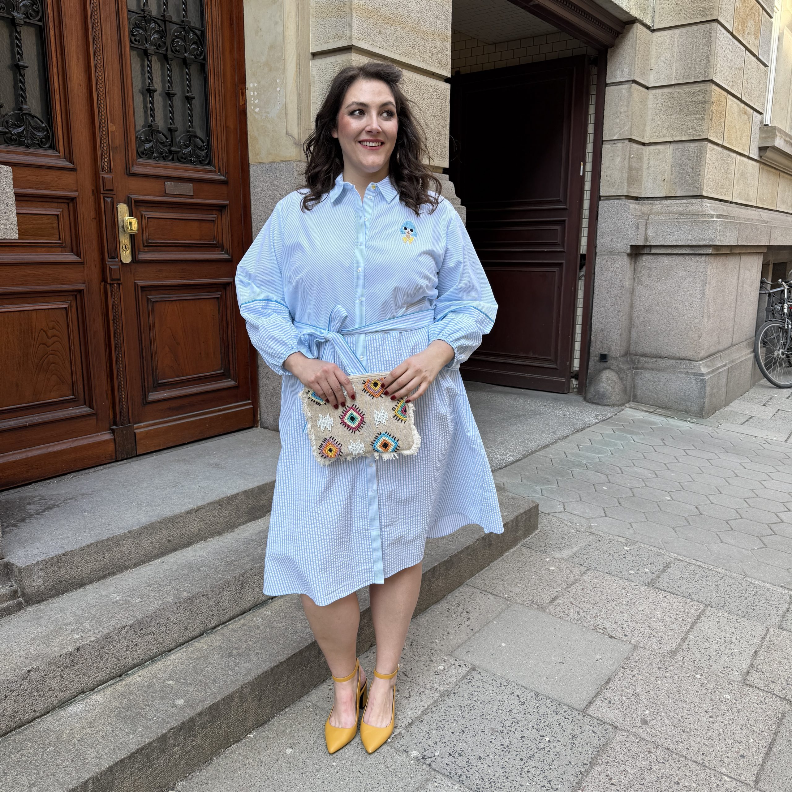 Große Größen in Bielefeld: Frau trägt hellblaues Oversize-Streifenkleid mit bunter Clutch und gelben Slingback-Pumps