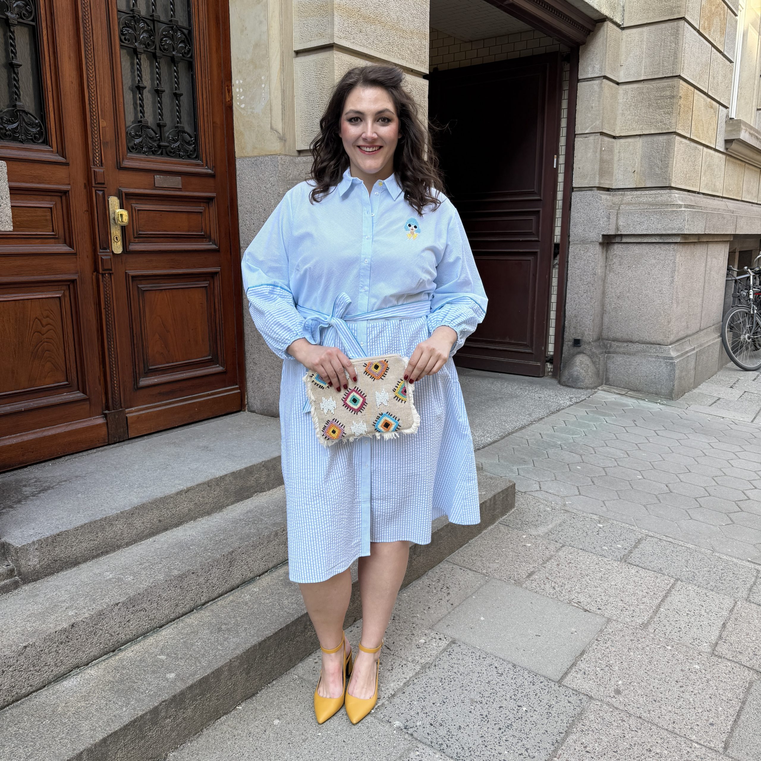 Große Größen in Braunschweig: Frau trägt hellblaues Streifenkleid mit bunter Clutch und gelben Slingback-Pumps