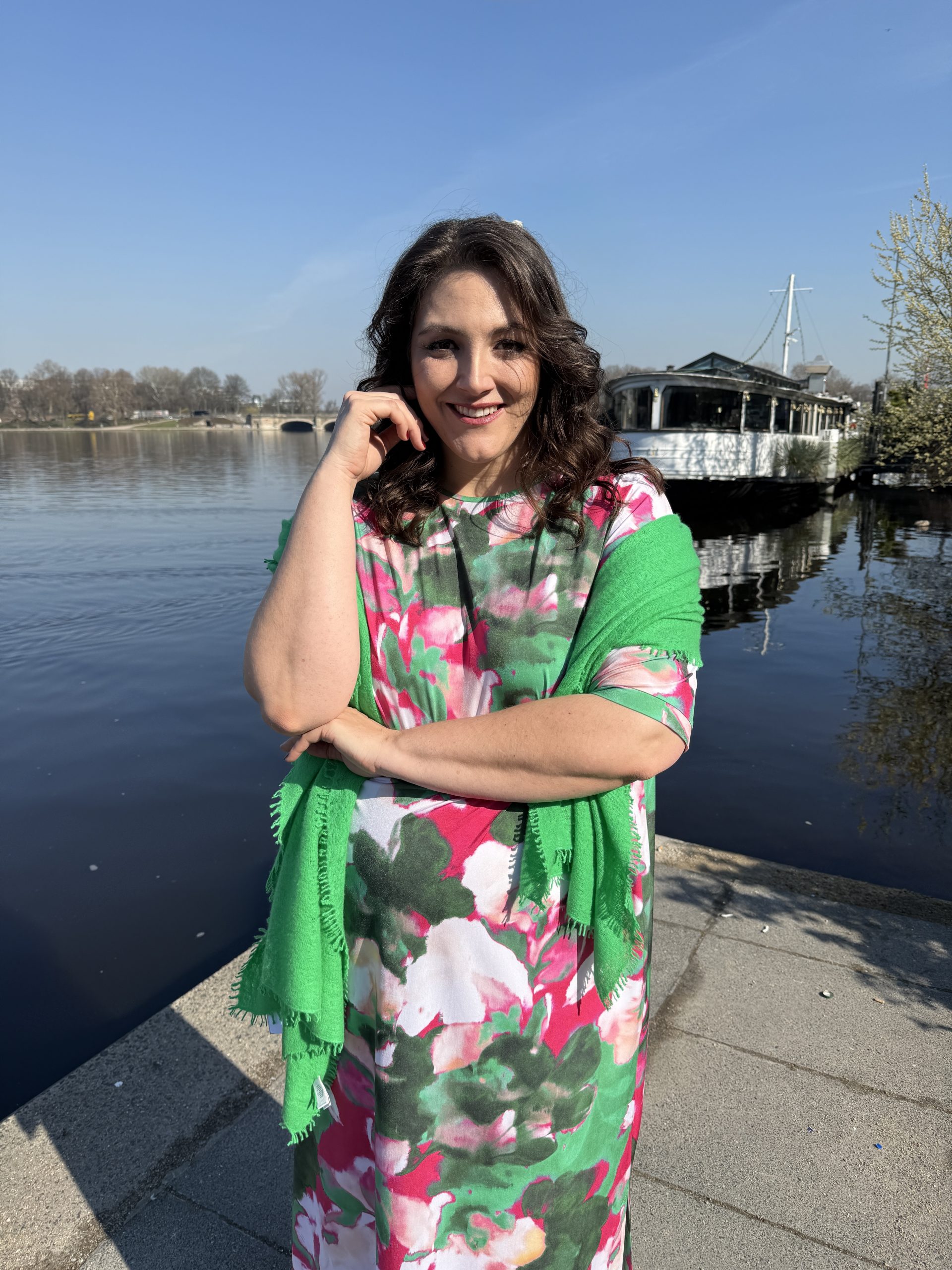Große Größen in Oldenburg: Frau trägt buntes Blumenkleid mit grünem Schal am Wasser im Frühling