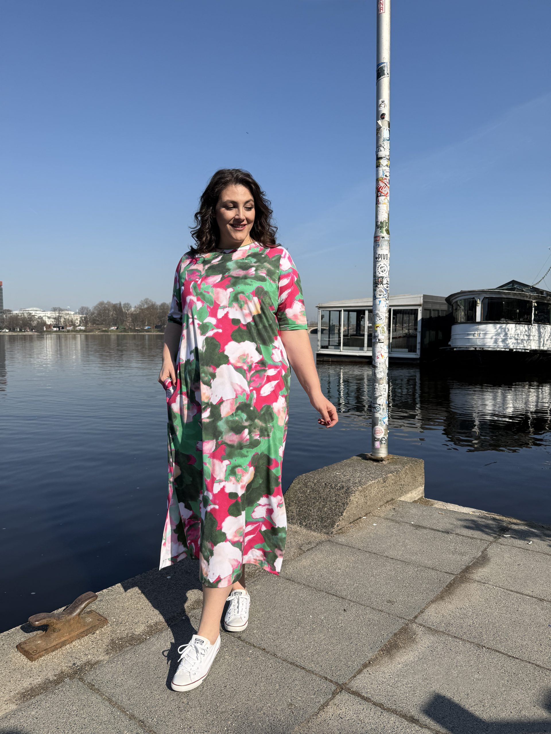 Mode für Mollige in Leipzig: Frau trägt langes pink-grünes Blumenkleid mit weißen Sneakers am Wasser