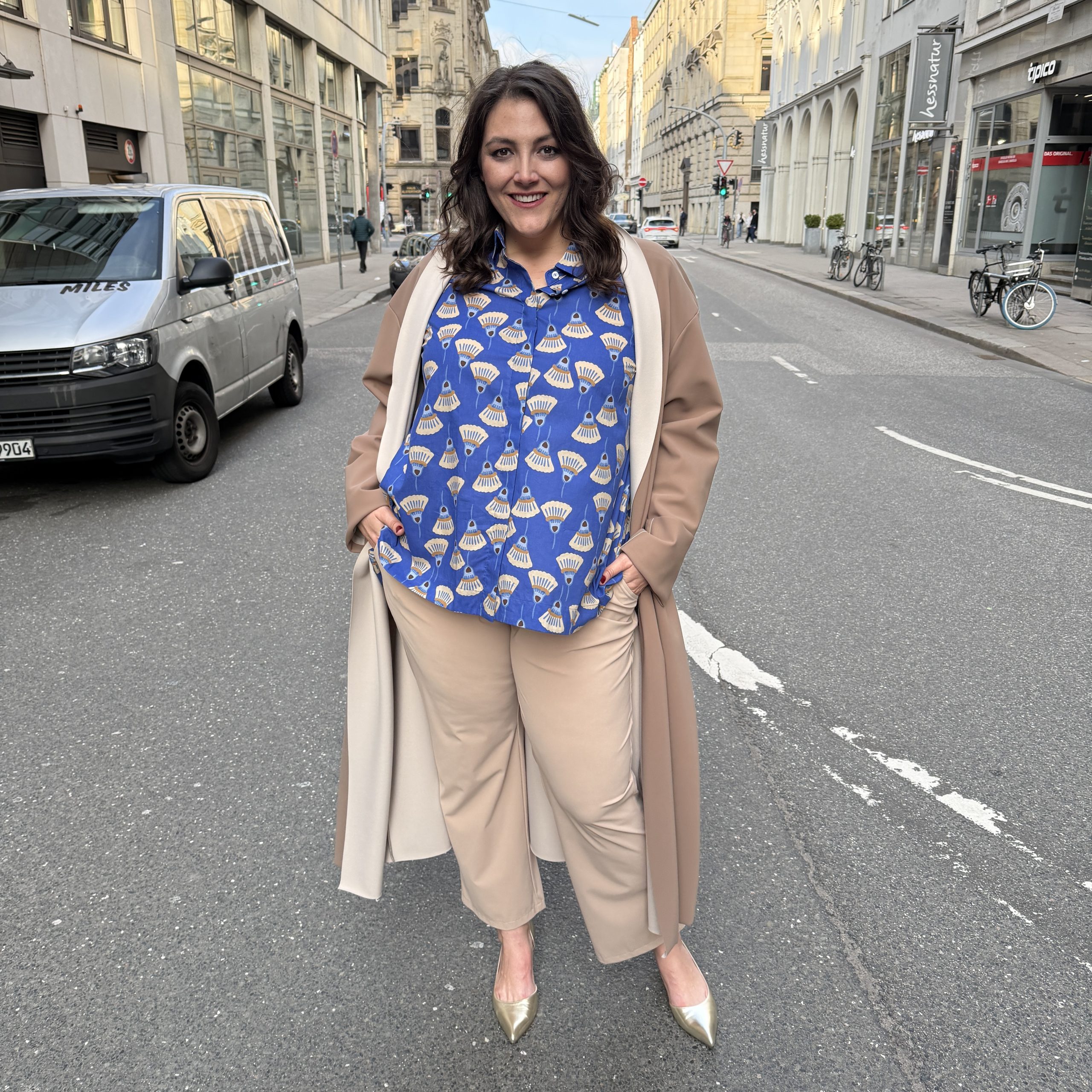 Große Größen in Leipzig: Frau trägt blaue Musterbluse mit beigem Longcoat und goldenen Pumps im Streetstyle
