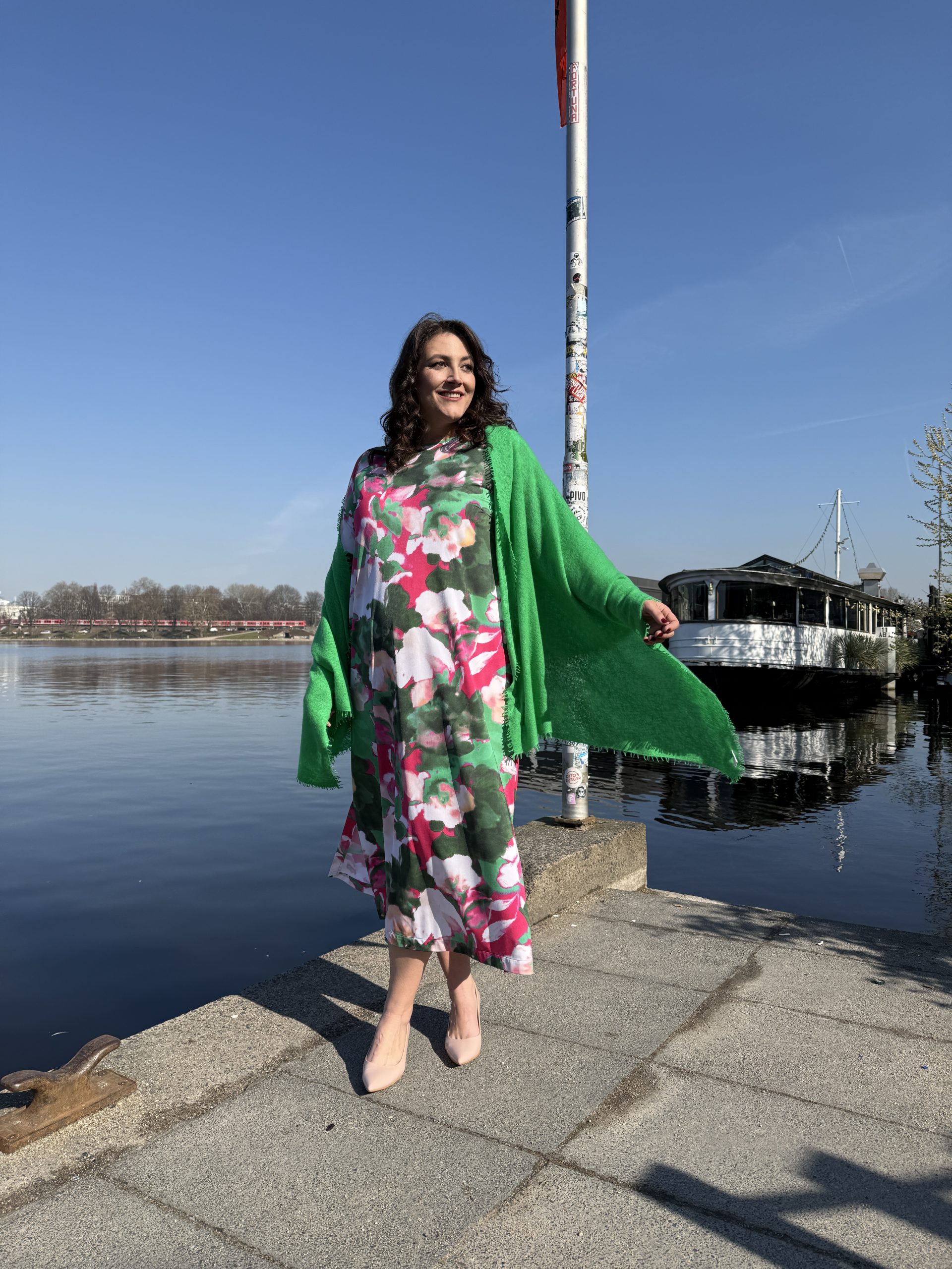 Mode für Mollige in Berlin: Frau trägt buntes Blumenkleid mit grünem Schal am Wasser