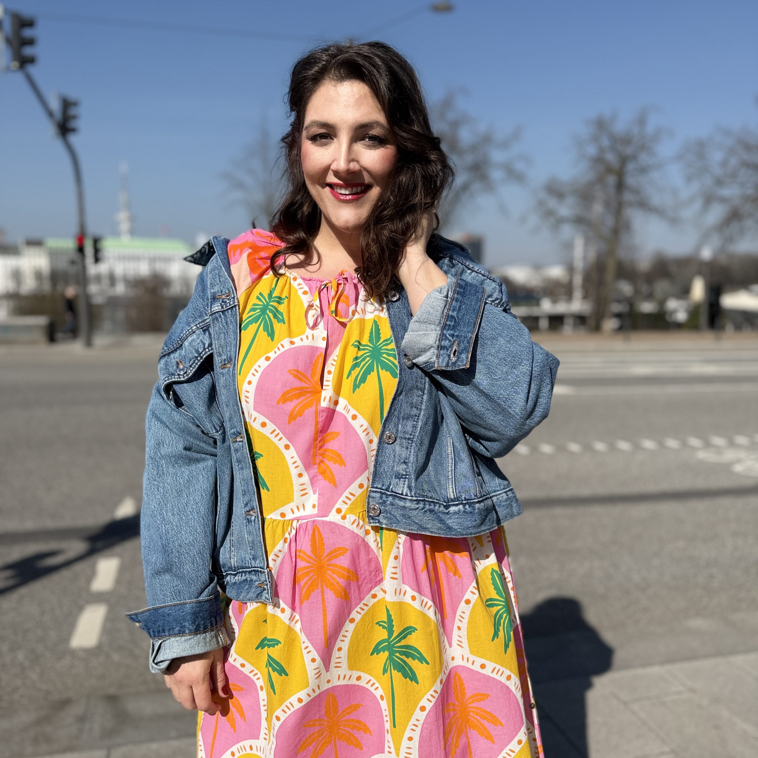 Mode für mollige Damen in München: Frau trägt buntes Palmenkleid mit Jeansjacke im Streetstyle