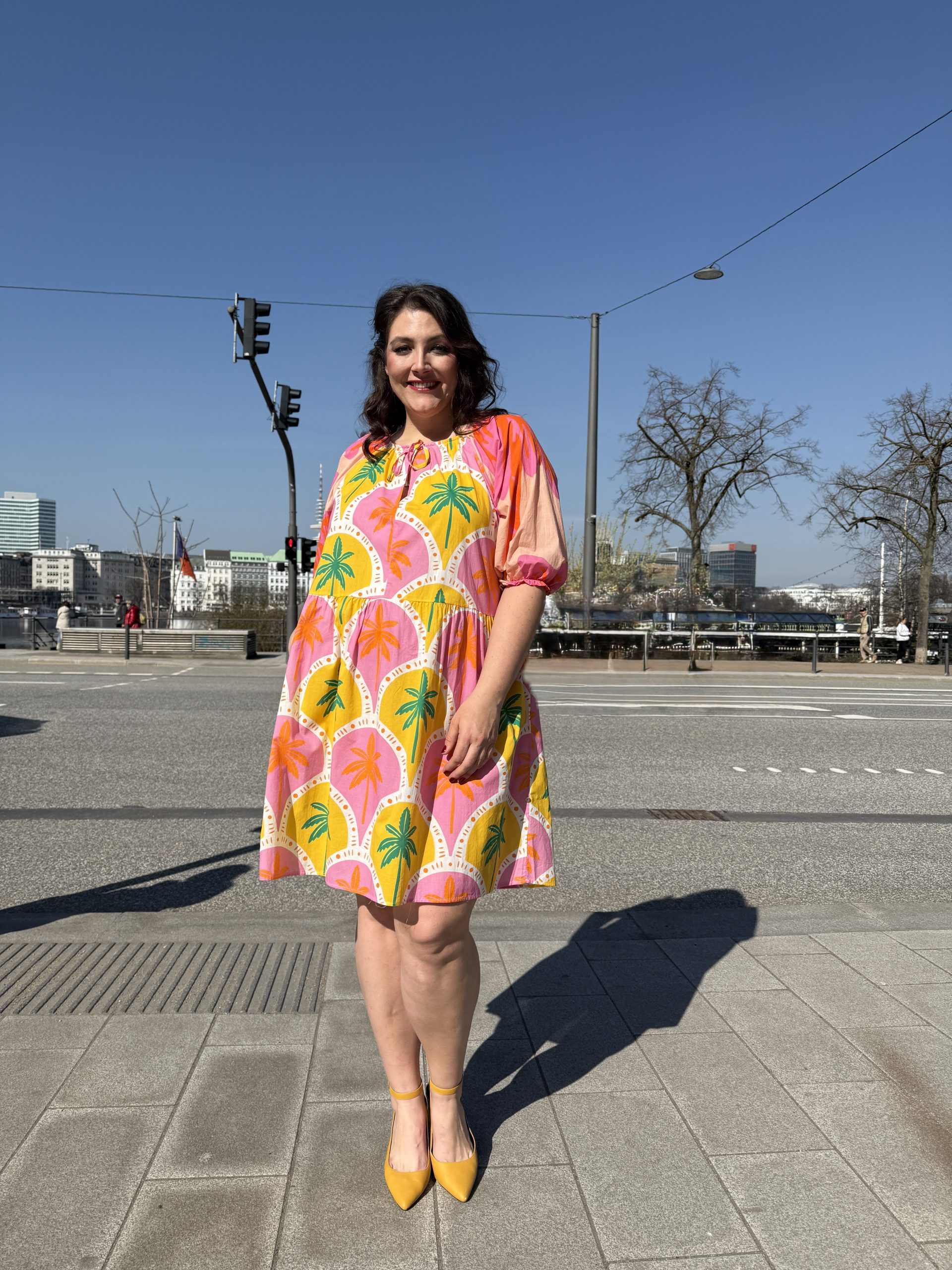 Mode für Mollige in Köln: Frau trägt buntes Sommerkleid mit Palmenprint und gelben Pumps am Rhein