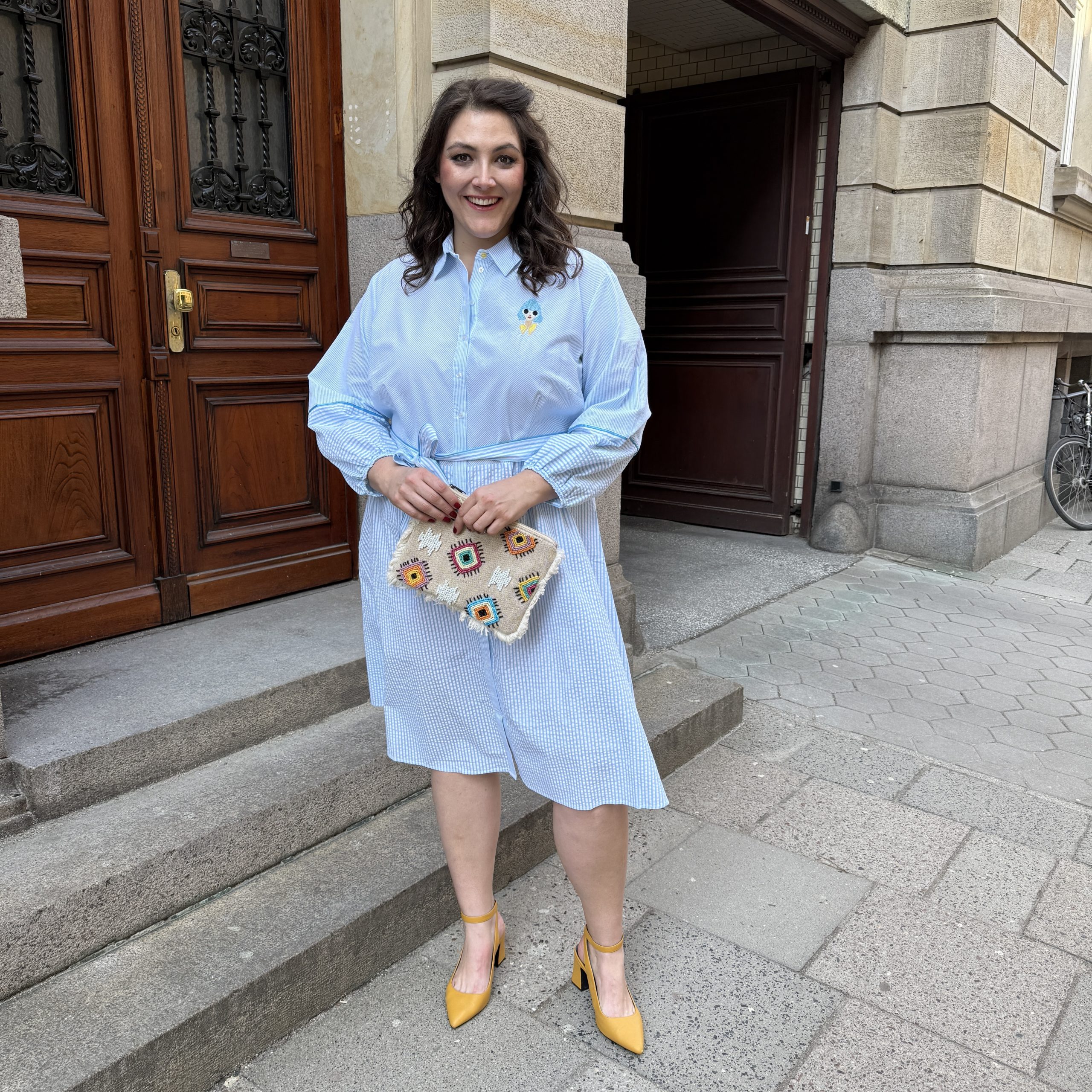 Mode für Mollige in München: Frau trägt hellblaues Streifenkleid mit bunter Clutch und gelben Pumps